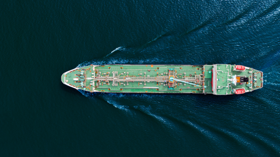 Aerial view of a large green cargo ship sailing on dark blue water leaving a wake behind.
