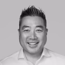 Smiling man with short spiked hair wearing a white collared shirt against a gray background.