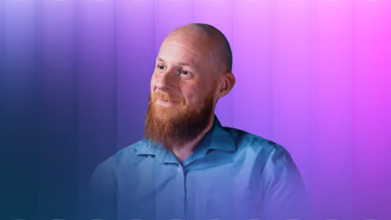 Smiling bald man with red beard wearing a blue shirt against a purple gradient background.