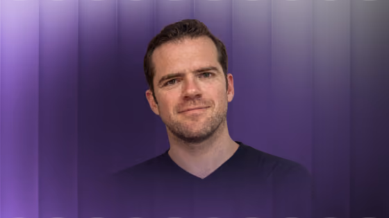 Man with short brown hair and light facial stubble wearing a black shirt against a purple gradient background.