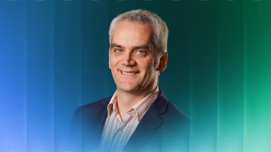 Smiling man with short gray hair wearing a dark blazer and striped shirt on blue-green background.