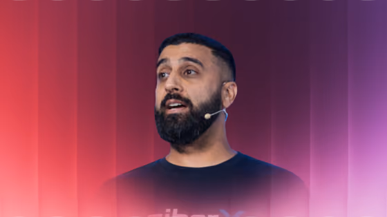 Bearded man speaking with a headset microphone against a red to purple gradient background.