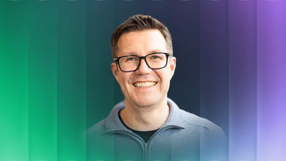 Smiling man wearing glasses and a gray zip-up shirt with a blurred green and purple background.