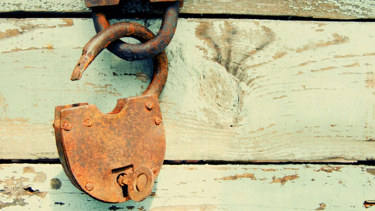 Close-up of an old rusty unlocked padlock hanging on a weathered pale wooden surface.