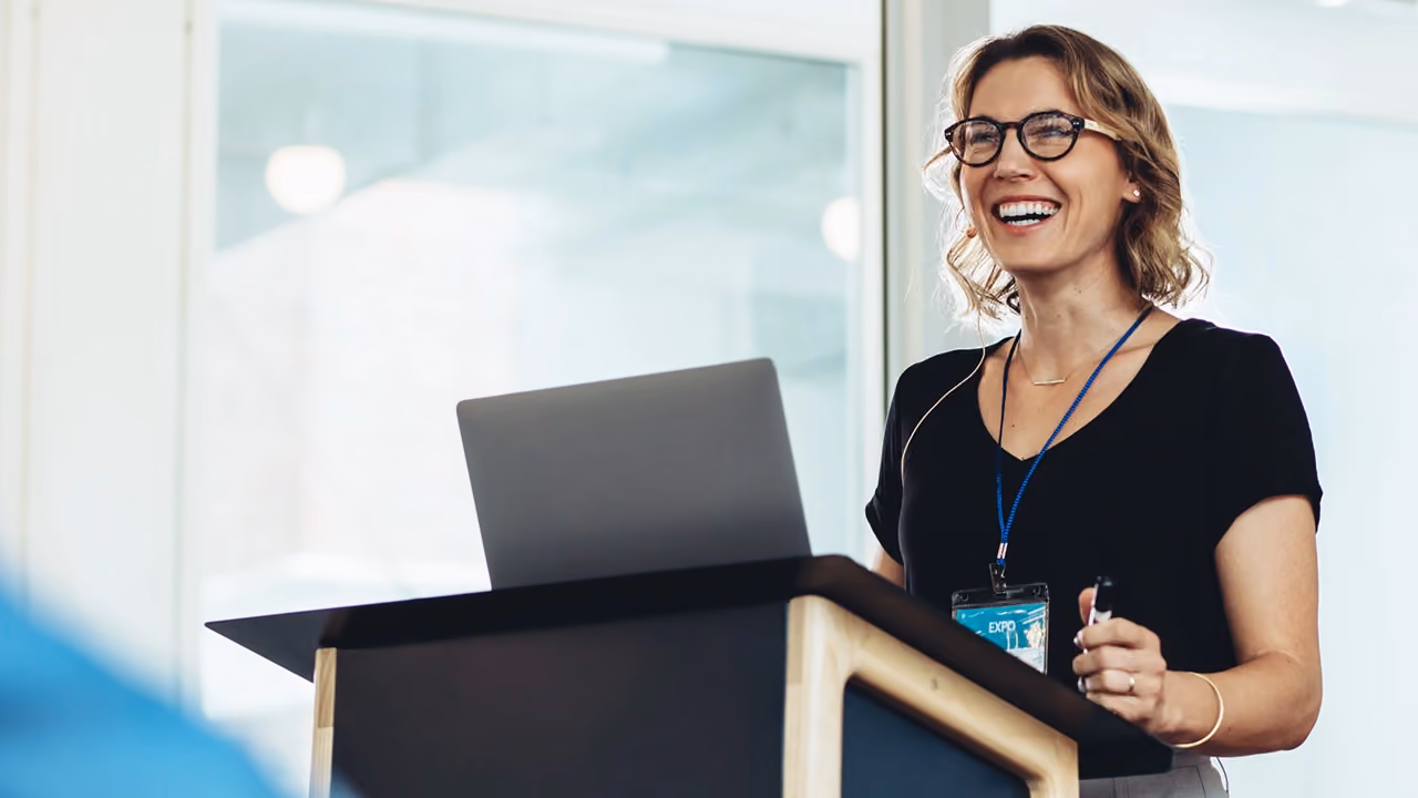 Smiling woman wearing glasses stands behind a podium with a laptop, holding a marker.