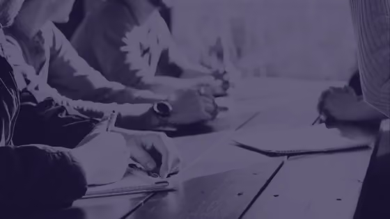 People writing notes on paper at a wooden table during a meeting or study session.