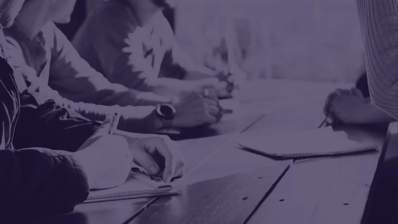 People sitting at a wooden table writing notes on notepads during a meeting or workshop.