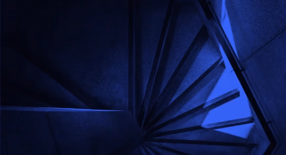 Dark blue spiral staircase viewed from above with sharp shadows and angles.