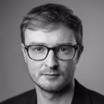 Black and white headshot of a man with short hair wearing glasses and a dark jacket, looking directly at the camera with a neutral expression.