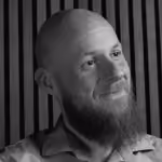 Portrait of a bald man with a full beard, wearing a collared shirt, smiling gently and looking to the side against a background of vertical dark wooden slats.