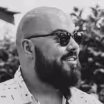 Black and white photo of a bald man with a beard and sunglasses, smiling and facing right with blurred foliage in the background.