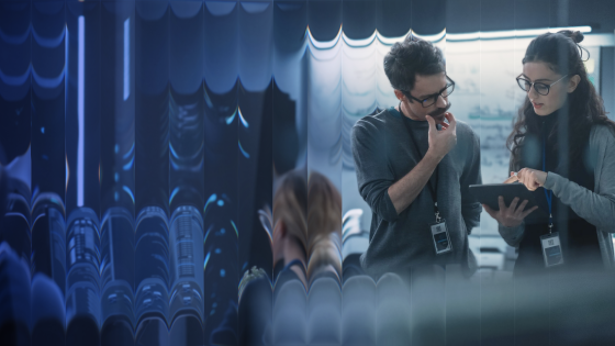 Two colleagues wearing badges discuss work using a tablet in a modern office setting.