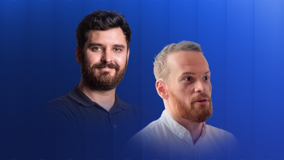 Two men standing side by side against a blue background, one with dark hair and beard wearing a black shirt, the other with light hair and beard wearing a white shirt, both looking slightly off to the side.