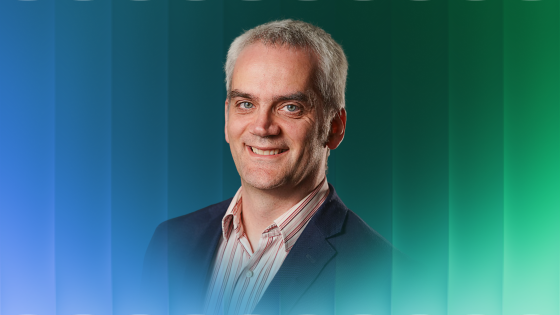 Smiling man with short gray hair wearing a dark blazer and striped shirt on blue-green background.