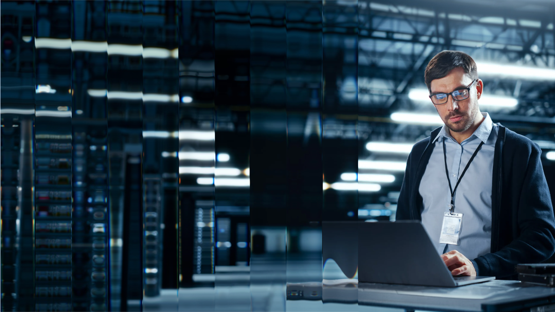 Man wearing glasses working on a laptop in a data center with server racks and blue lighting.