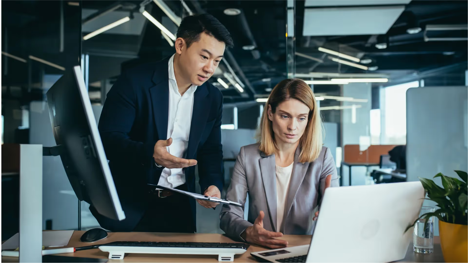 Man in suit showing clipboard to seated woman in blazer working on a laptop in office.