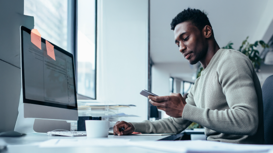 Man sitting at desk in front of computer, focused on using a smartphone in a bright office.