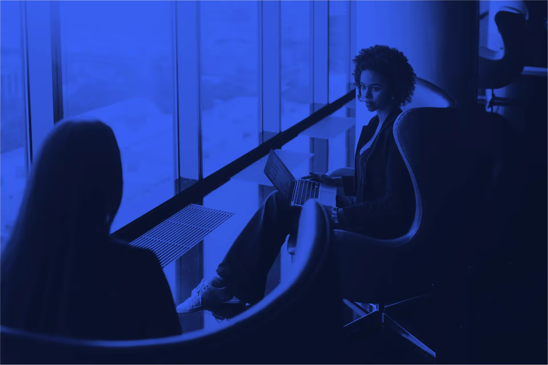 Two women sitting by large windows, one with a laptop showing a chart, in a modern office space.