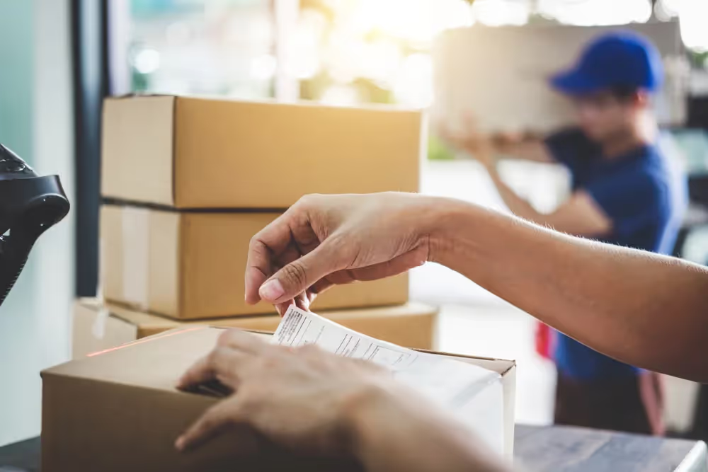 Person attaching a shipping label to a cardboard box with stacked boxes and a delivery person in the background.