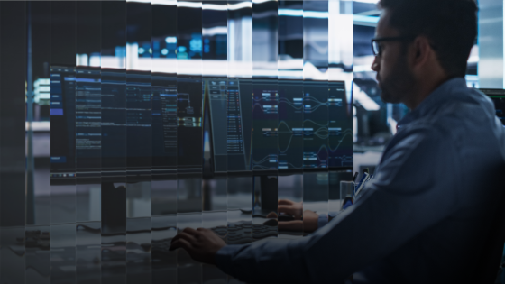 Man wearing glasses analyzing data on multiple computer monitors in a dimly lit room.