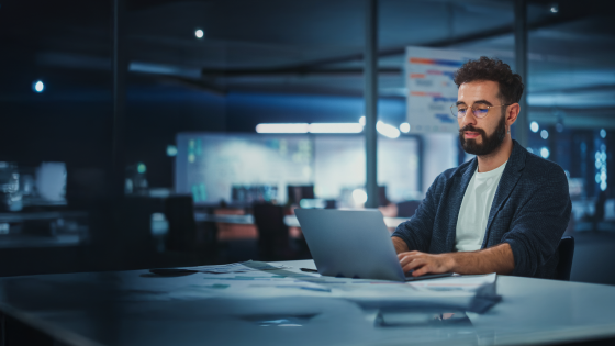 Man with glasses working on a laptop at a desk in a modern office space at night.