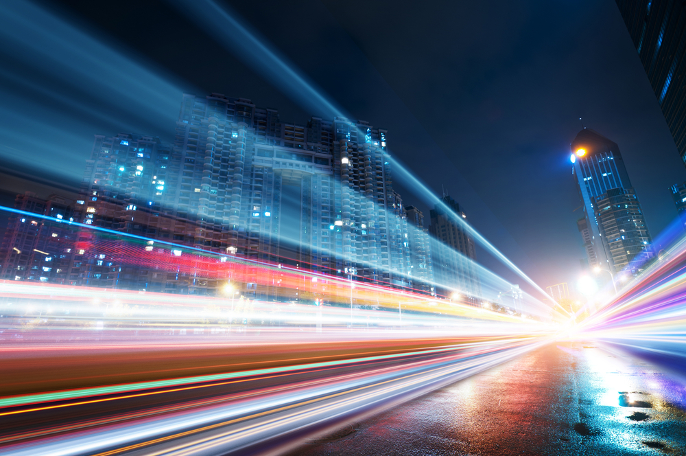Night cityscape with tall buildings and colorful light trails from fast-moving vehicles on wet street.