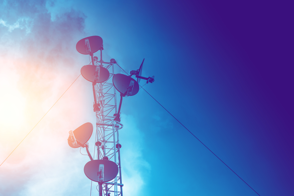 Telecommunication tower with multiple satellite dishes against a blue sky with sun flare.