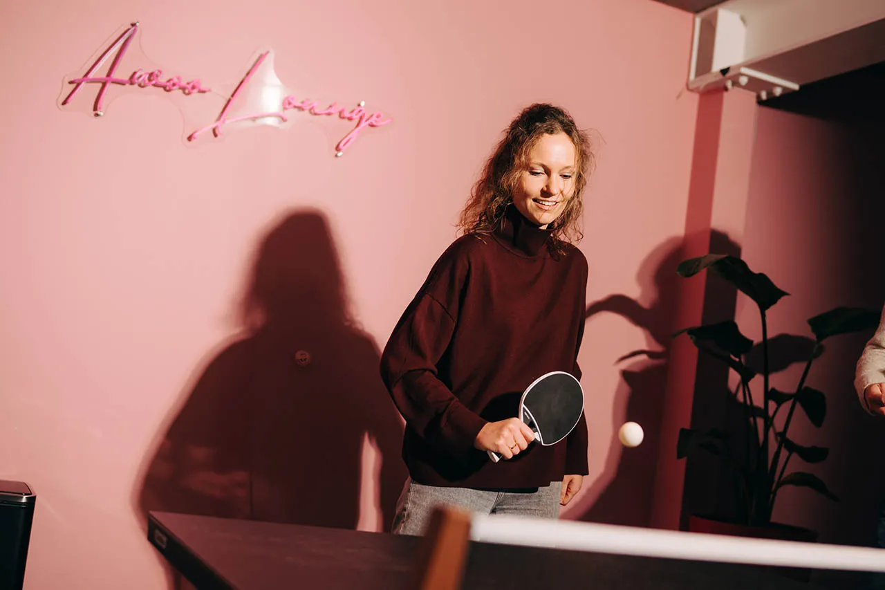Woman playing table tennis against a pink wall lit by a cursive neon sign