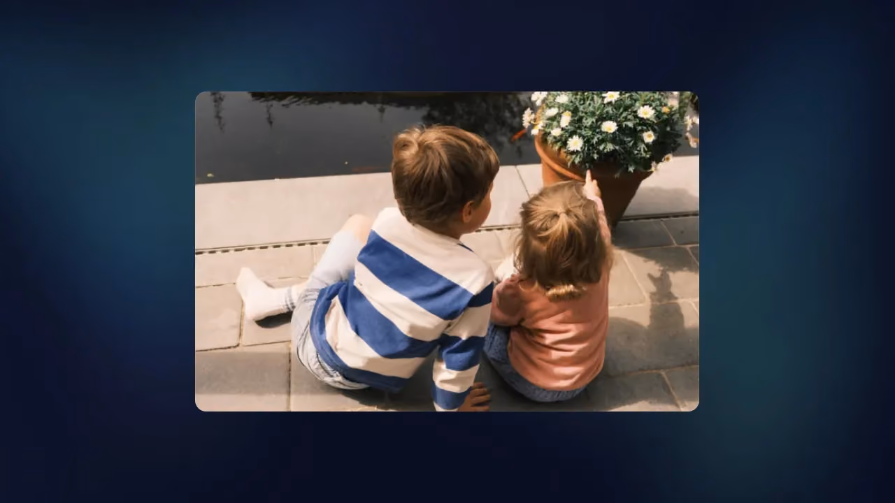 Two young children sitting on a quay by the water, pointing at a pot of daisies