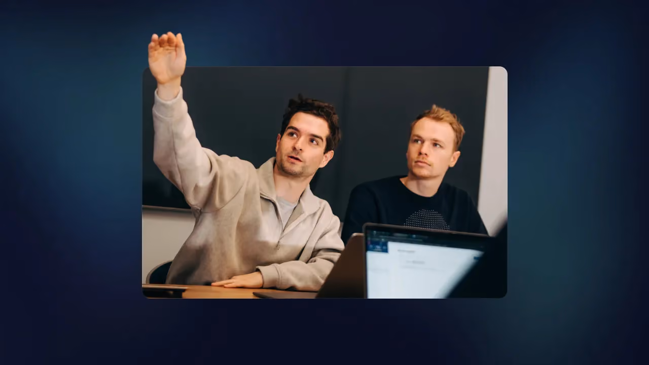 Two men at a desk, one raising his hand to ask a question during a session