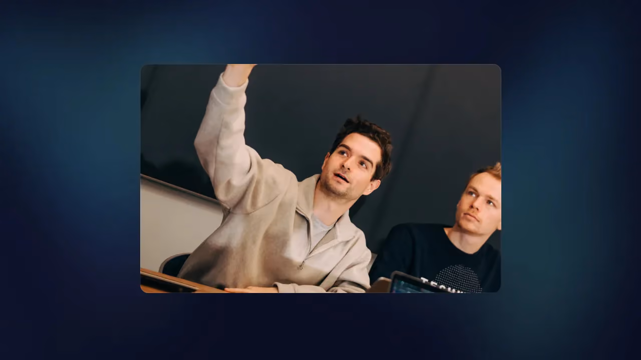 Two colleagues at a desk reviewing code on a large monitor, one pointing at the screen