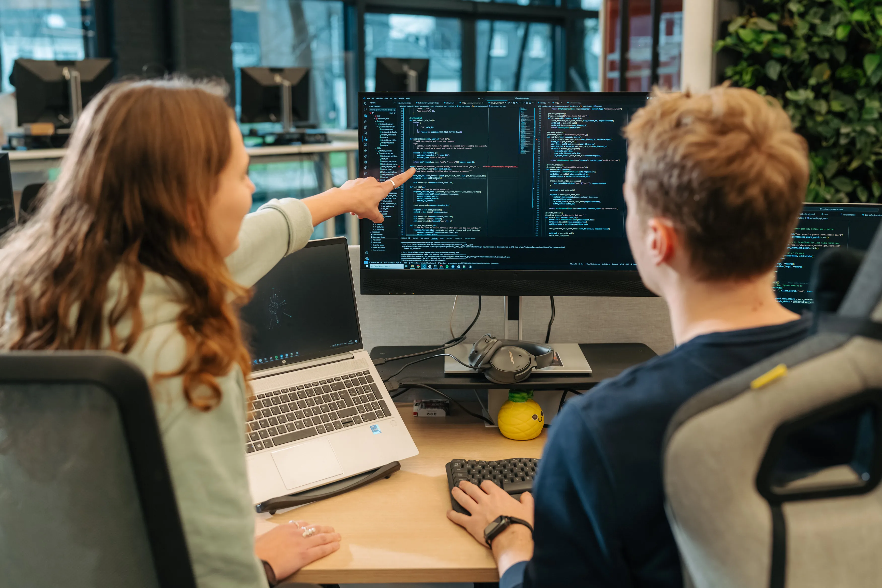 Two colleagues at a desk reviewing code on a large monitor, one pointing at the screen