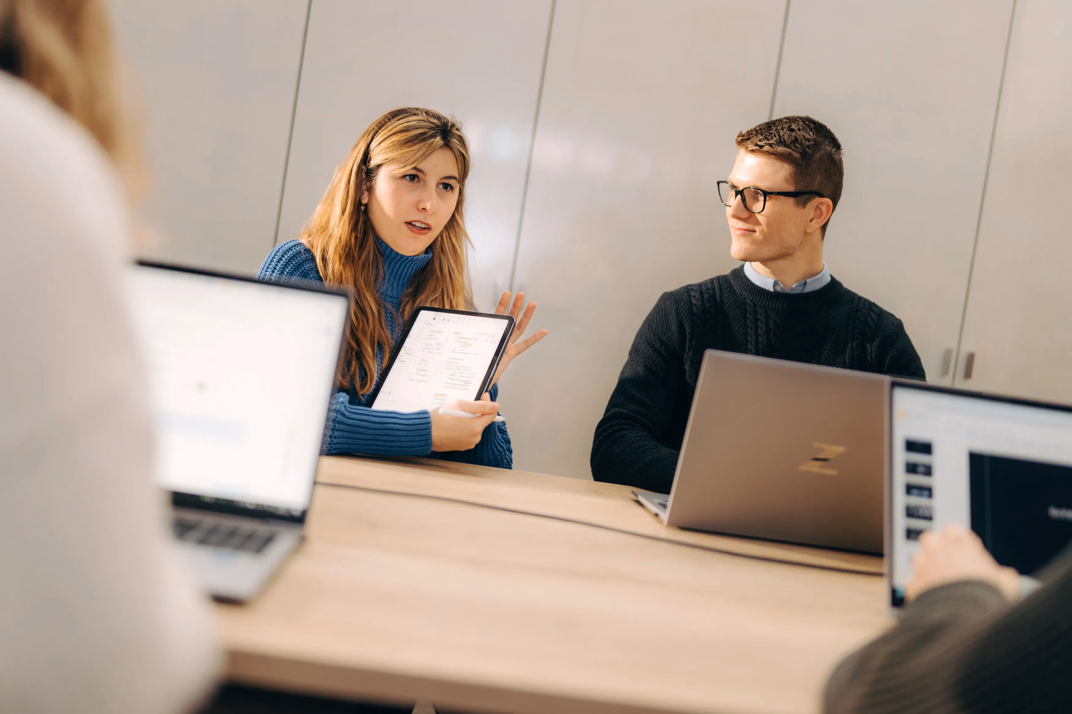 Woman holding a tablet speaking to colleagues around a meeting table covered in laptops