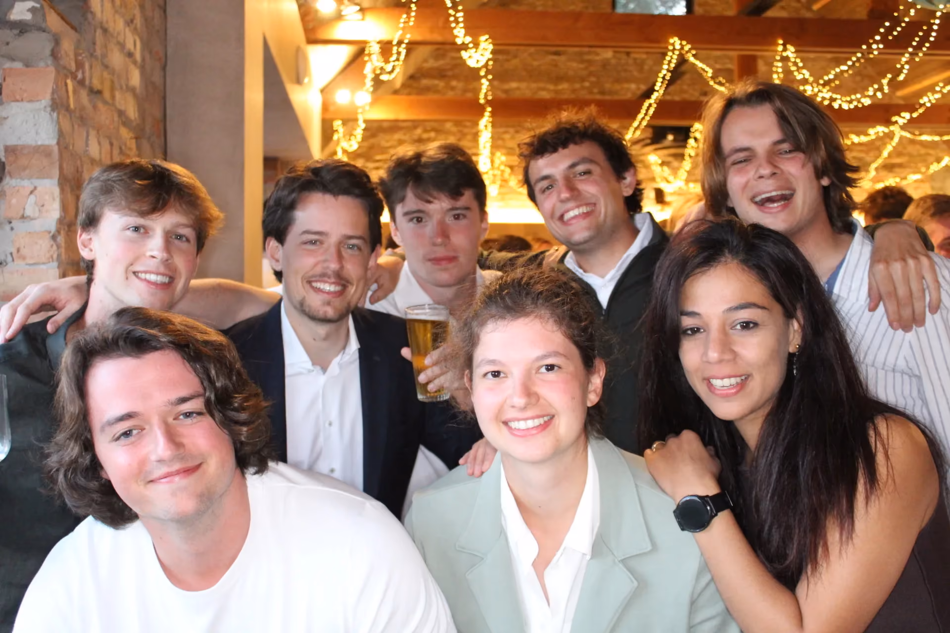 Group photo of eight smiling colleagues at an evening party under warm string lights