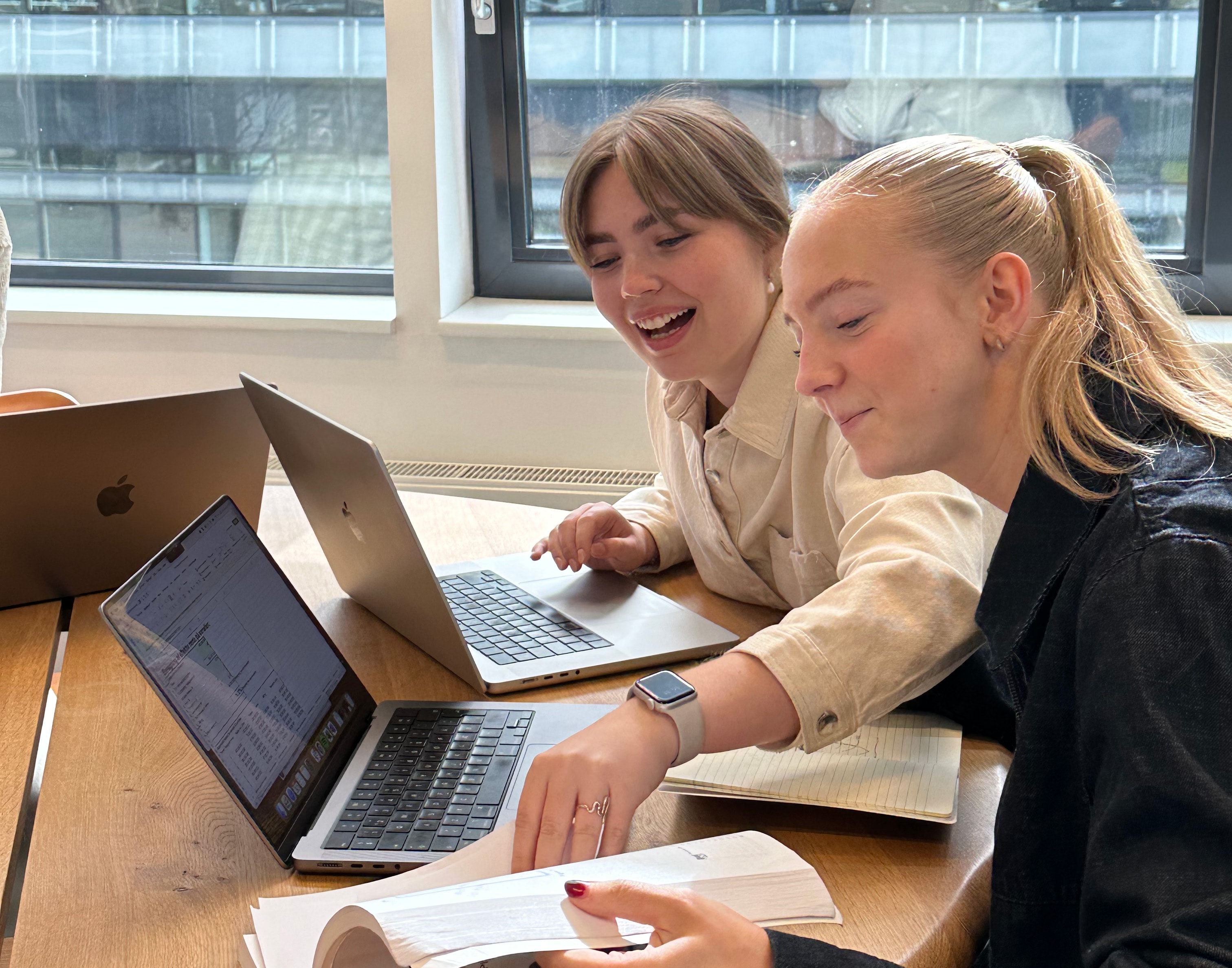 Two young women working together at a table with laptops and papers, smiling and engaged.
