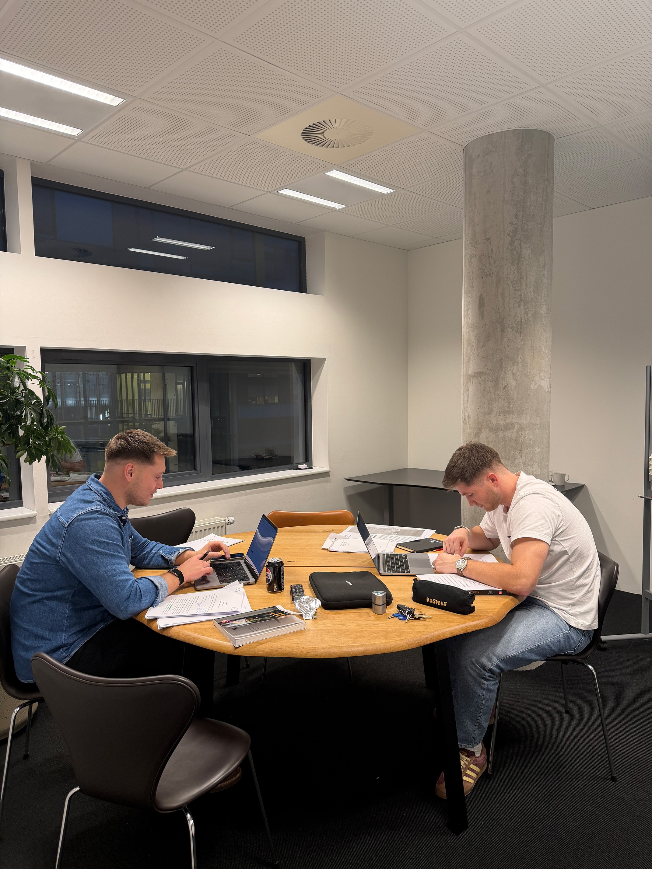 Two young men working on laptops and paperwork at a round wooden table in a modern office room.