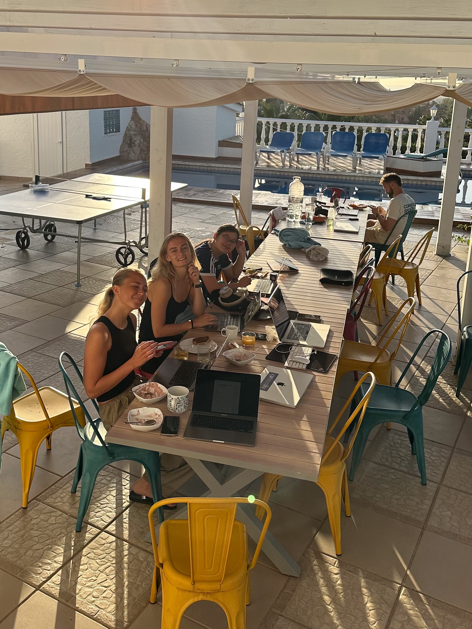 Group of four young adults working on laptops and eating at a long outdoor table near a pool under a shaded patio.