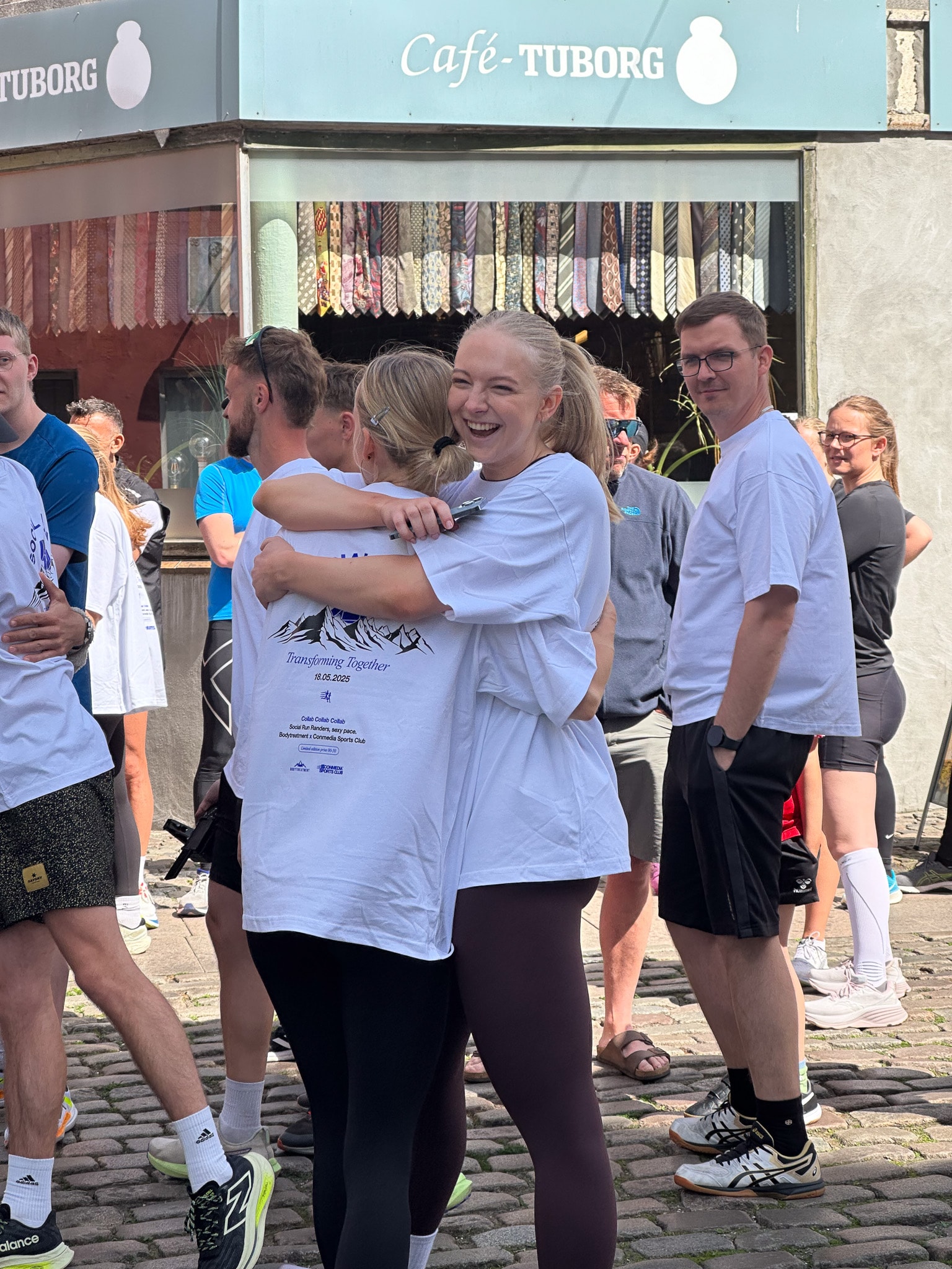 Two young women in white event t-shirts hug and smile outdoors on a cobblestone street in front of Café Tuborg, while others stand and socialize in the background.