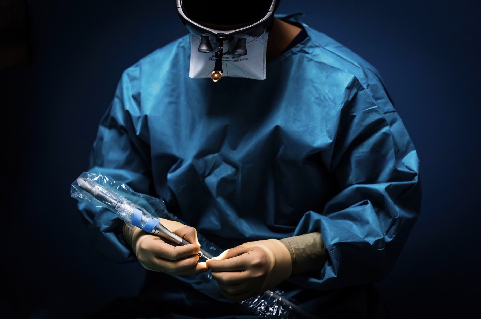 Gloved hands of a surgeon in blue gown working with surgical tools under focused light.