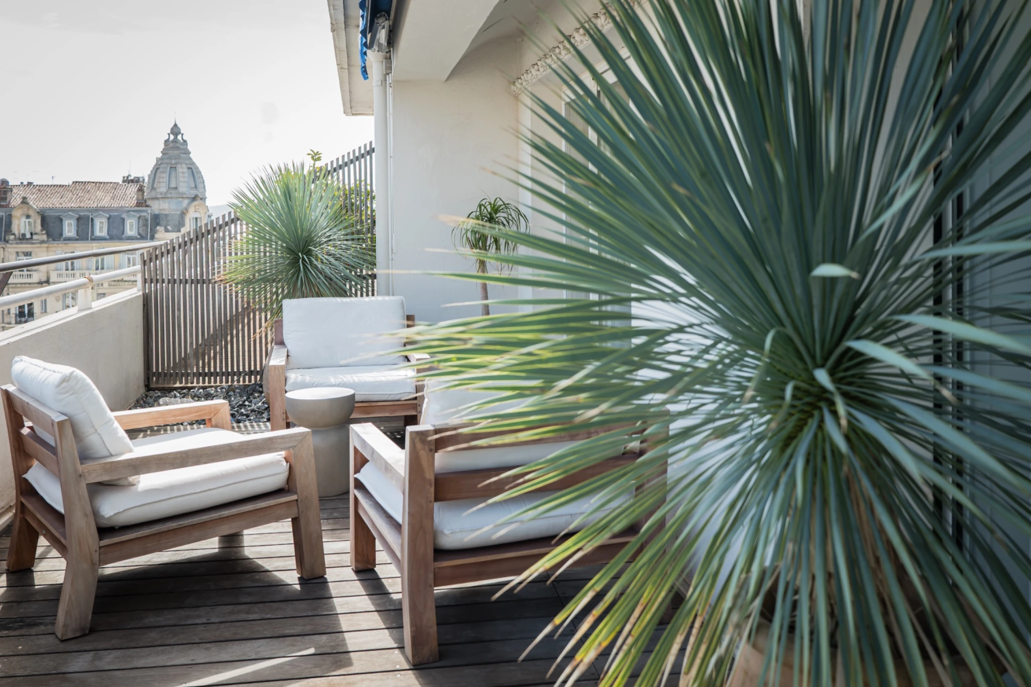 Balcony seating with wooden chairs, white cushions, small table, and large spiky plants.