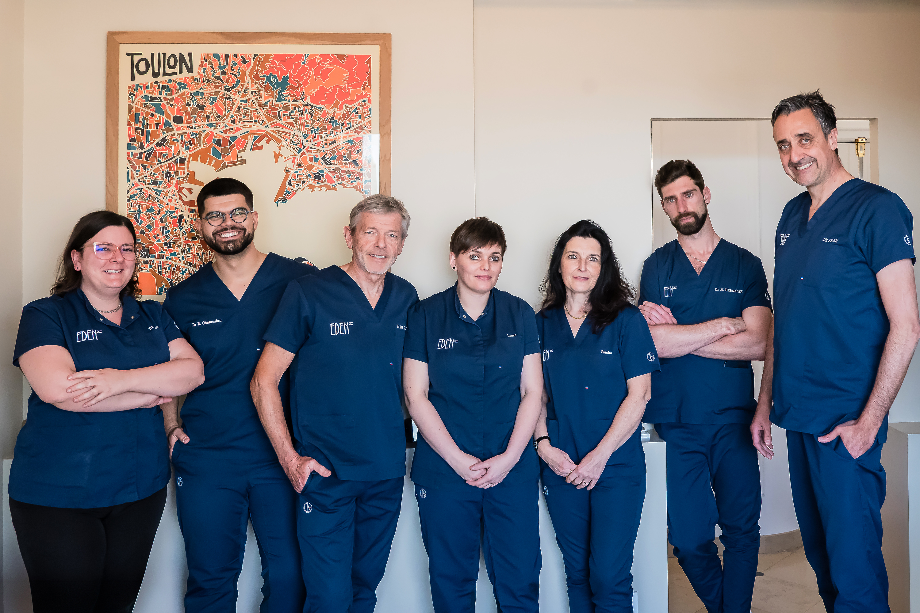 Seven smiling medical professionals in blue scrubs standing indoors with a colorful map of Toulon on the wall.