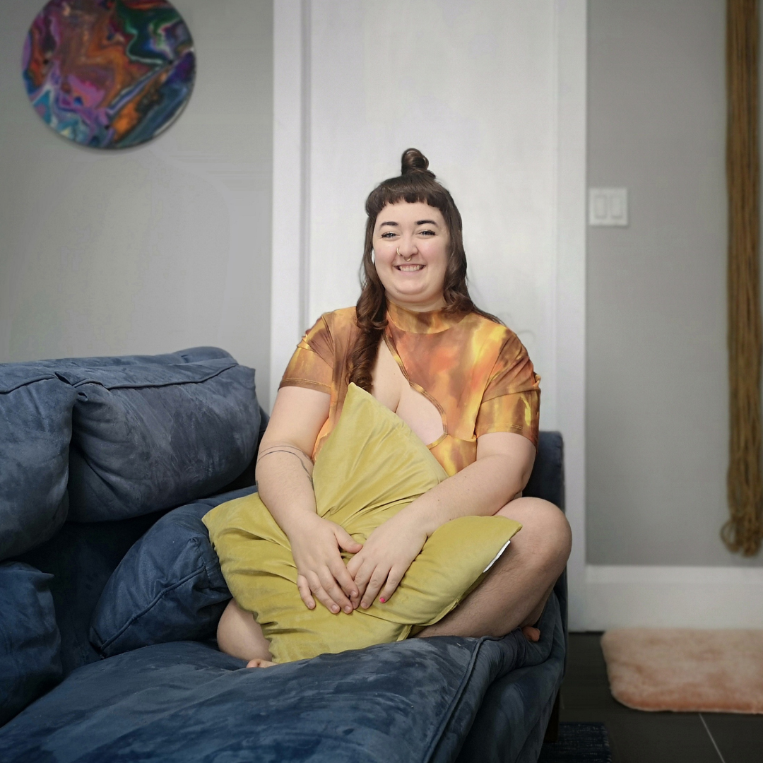 Smiling person sitting cross-legged on a blue couch holding a yellow pillow, with abstract wall art in the background. This is Linden.