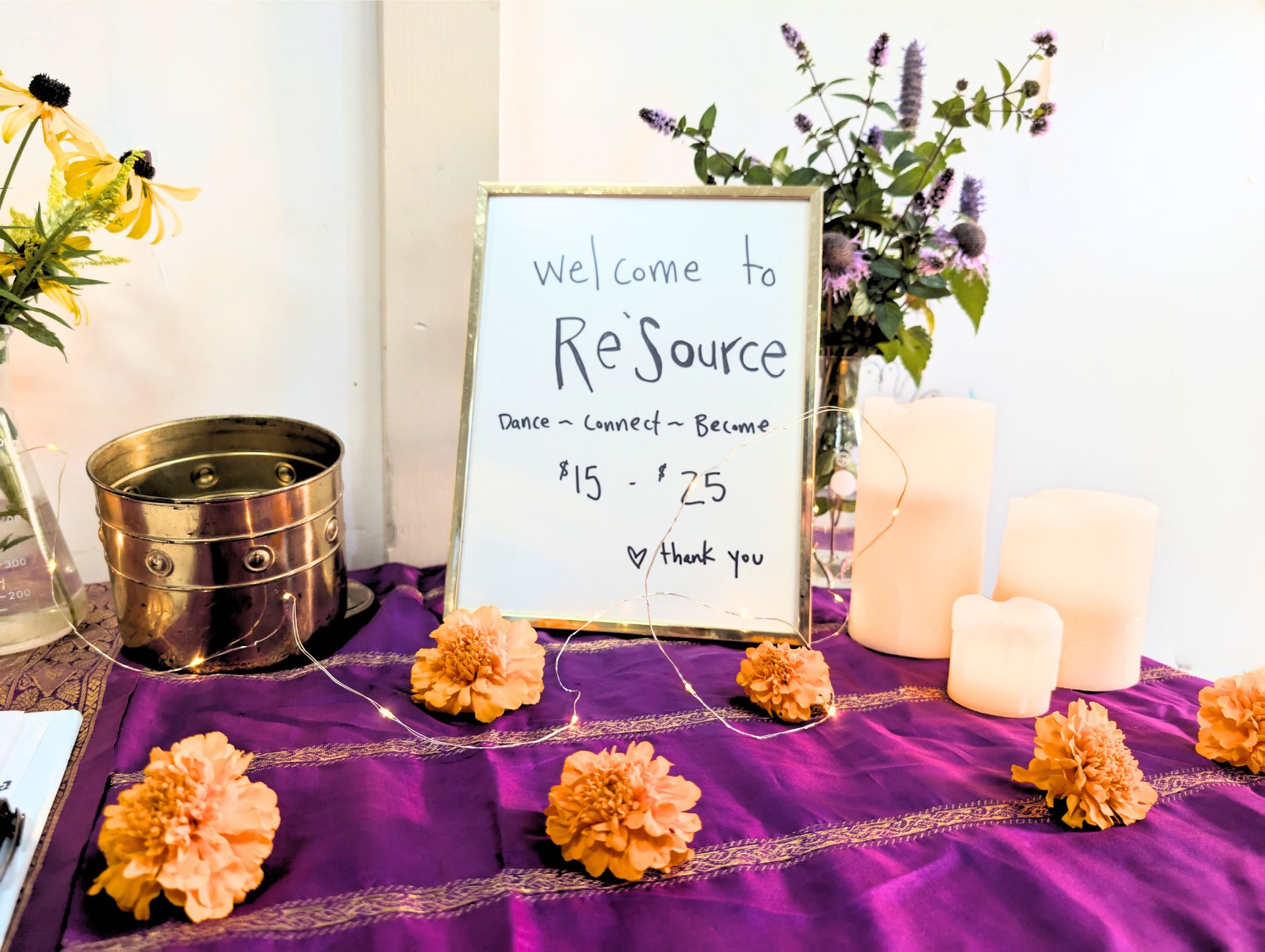 Table with a purple cloth displays a framed sign reading 'welcome to Re'Source Dance ~ Connect ~ Become $15 - $25 thank you,' surrounded by orange marigold flowers, LED string lights, a brass container, white candles, and vases with yellow and purple flowers.