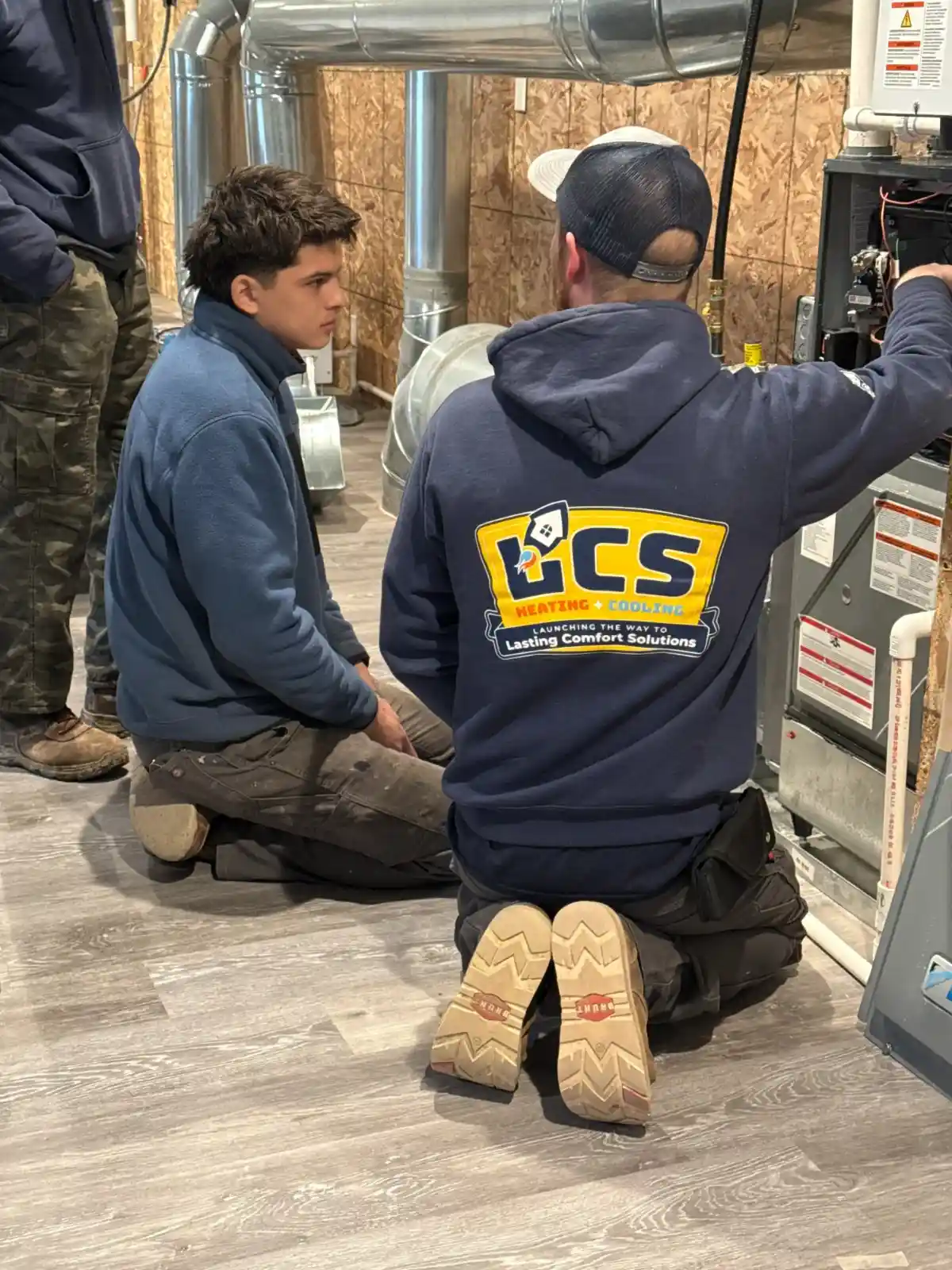 Two technicians, one in a hoodie with a logo, kneel on a wooden floor, focused on repairing a furnace. 