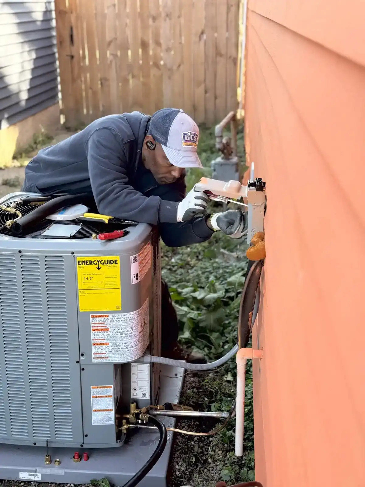 A technician in a cap and gloves works on an air conditioning unit next to an orange wall.