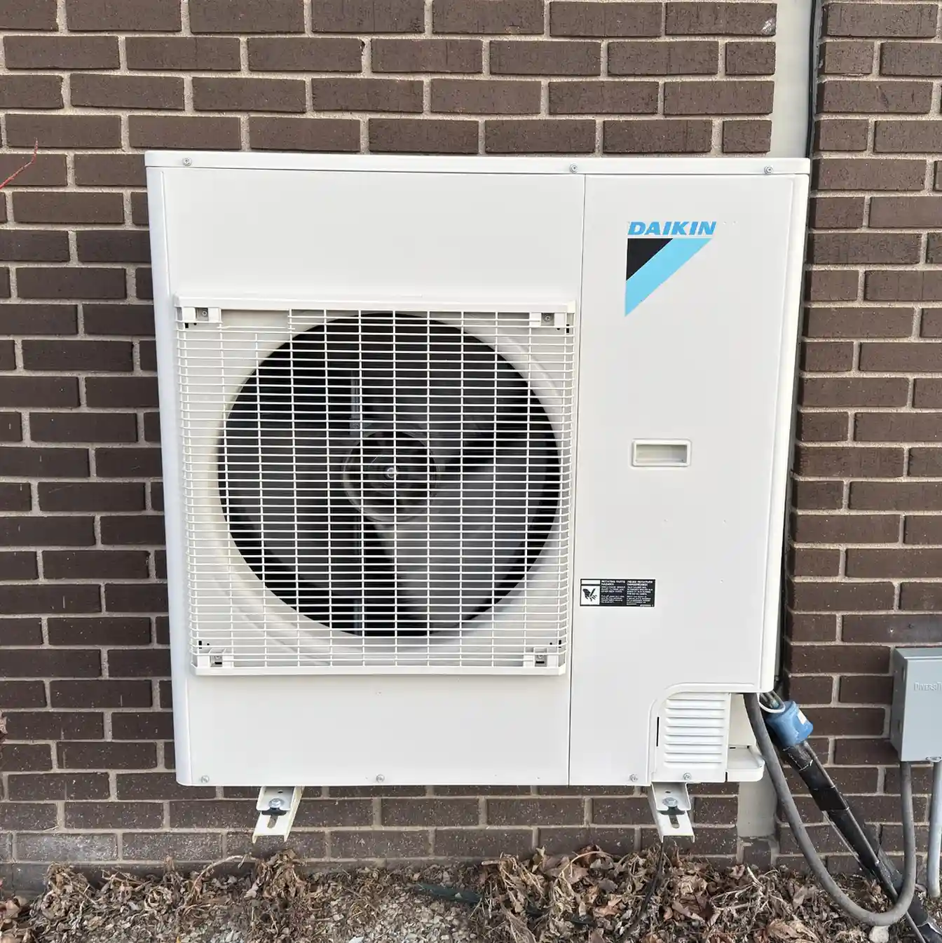Outdoor air conditioning unit mounted on a brick wall, featuring a large fan behind a protective grille. 