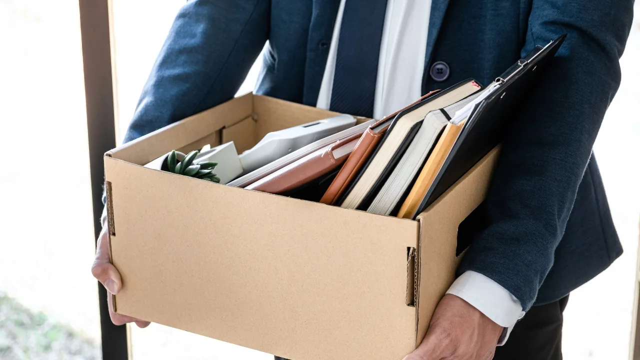 Homme en costume tenant une boîte en carton avec des affaires de bureau, évoquant un départ