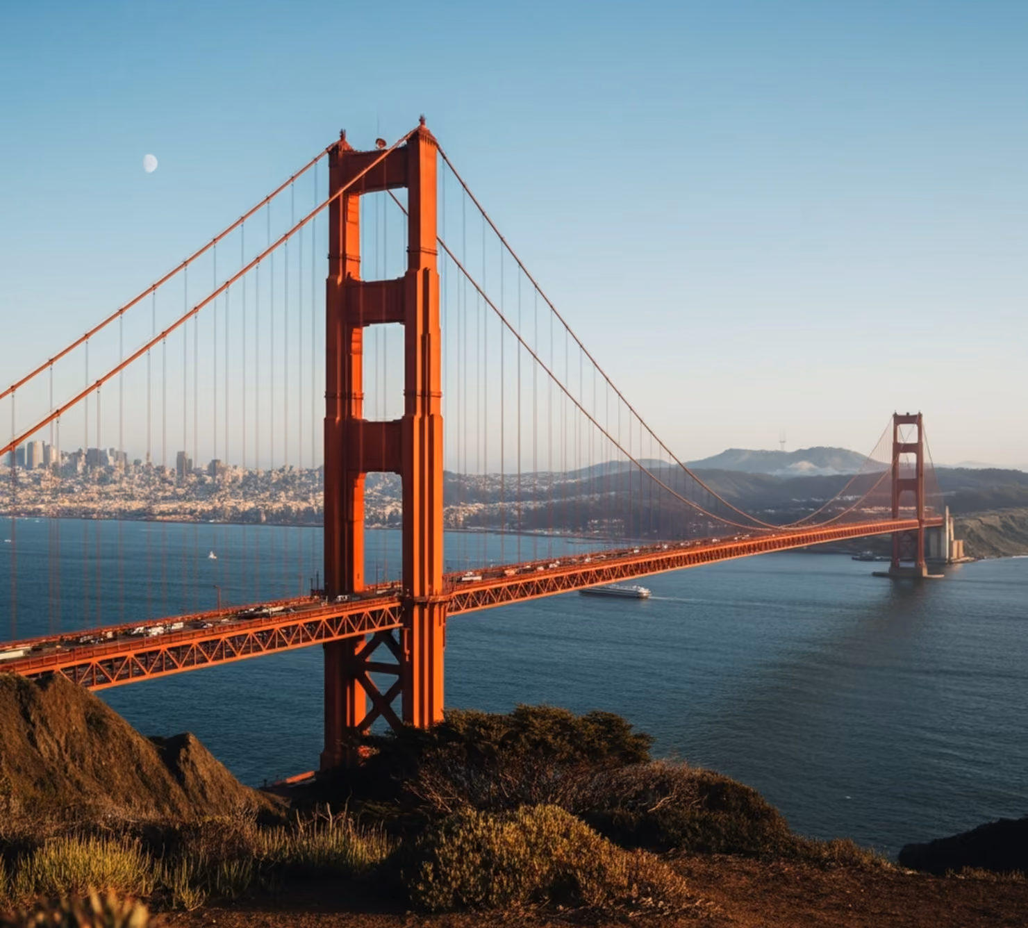 The red Golden Gate Bridge spanning across dark blue waters toward the San Francisco skyline under a clear sky with a faint moon visible.