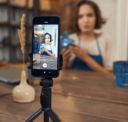 Woman recording a video on a smartphone mounted on a tripod while holding a small blue object.