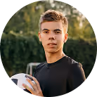 Young man in black shirt holding a white volleyball outdoors with green trees in background.